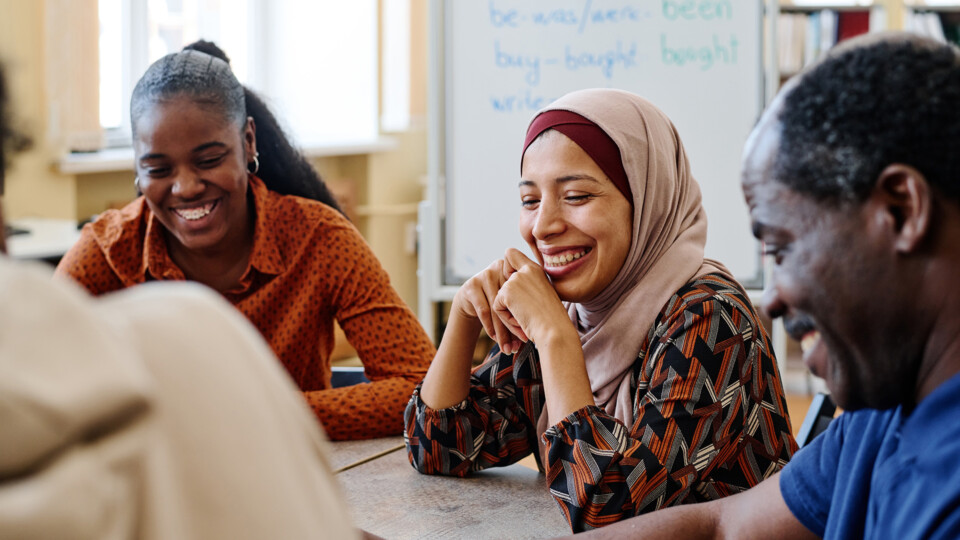 Group of modern immigrants sitting at table having fun laughing at something funny during english lesson