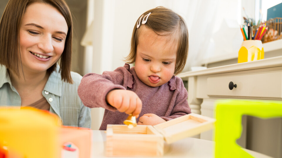 Portrait view of the happy woman playing with her little daughter with down syndrome at the table. Woman having fun with her daughter. Stock photo